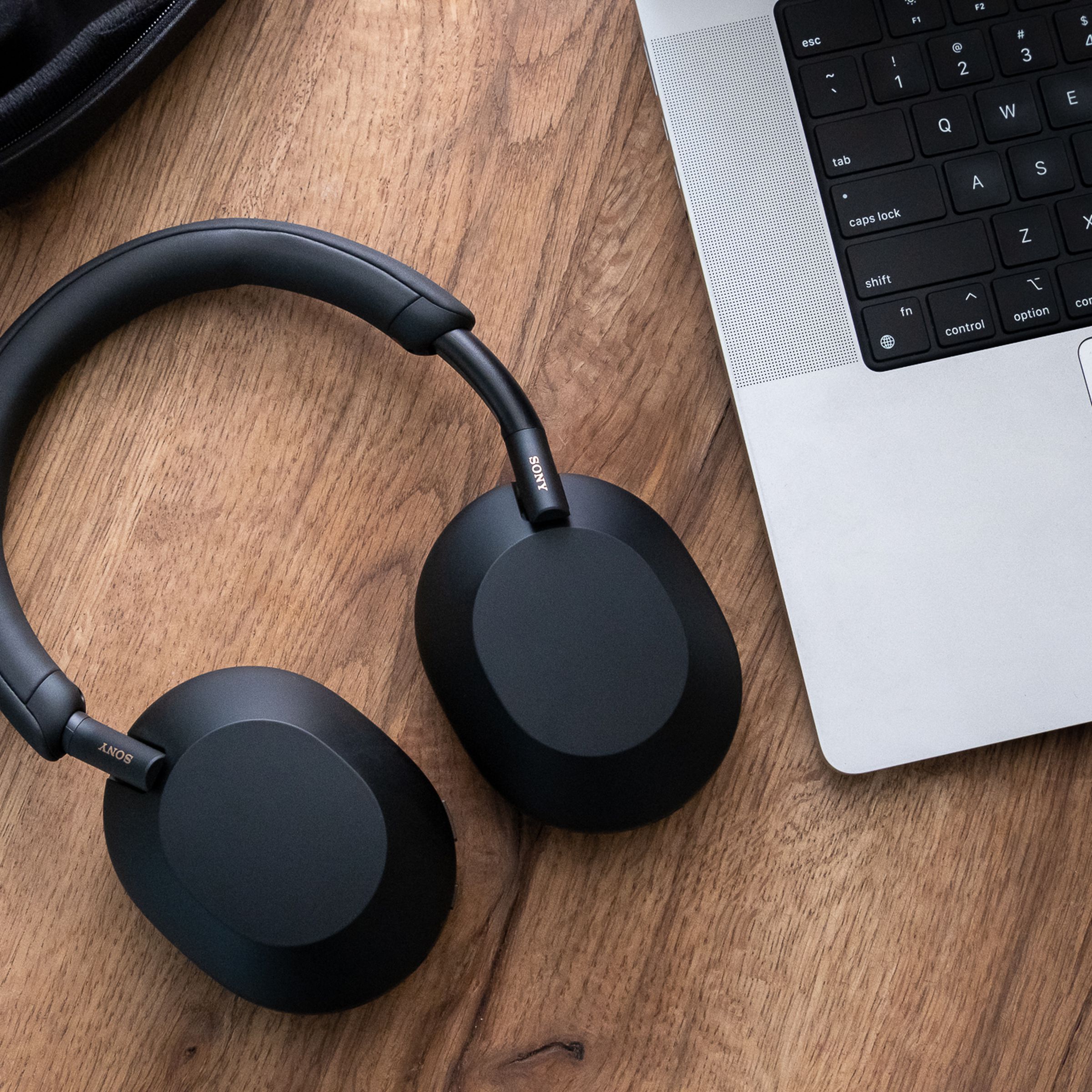 A pair of Sony WH-1000XM5 headphones, in black, resting flat on a wood table beside a laptop.