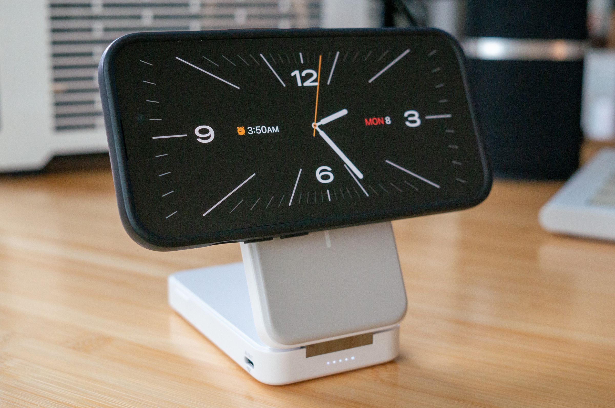 An iPhone in StandBy mode, showing an analog clock face, magnetically attached to a white and grey folding charging stand. The stand is thick, and has a silver hinge and a row of white LEDs indicating battery charge level.