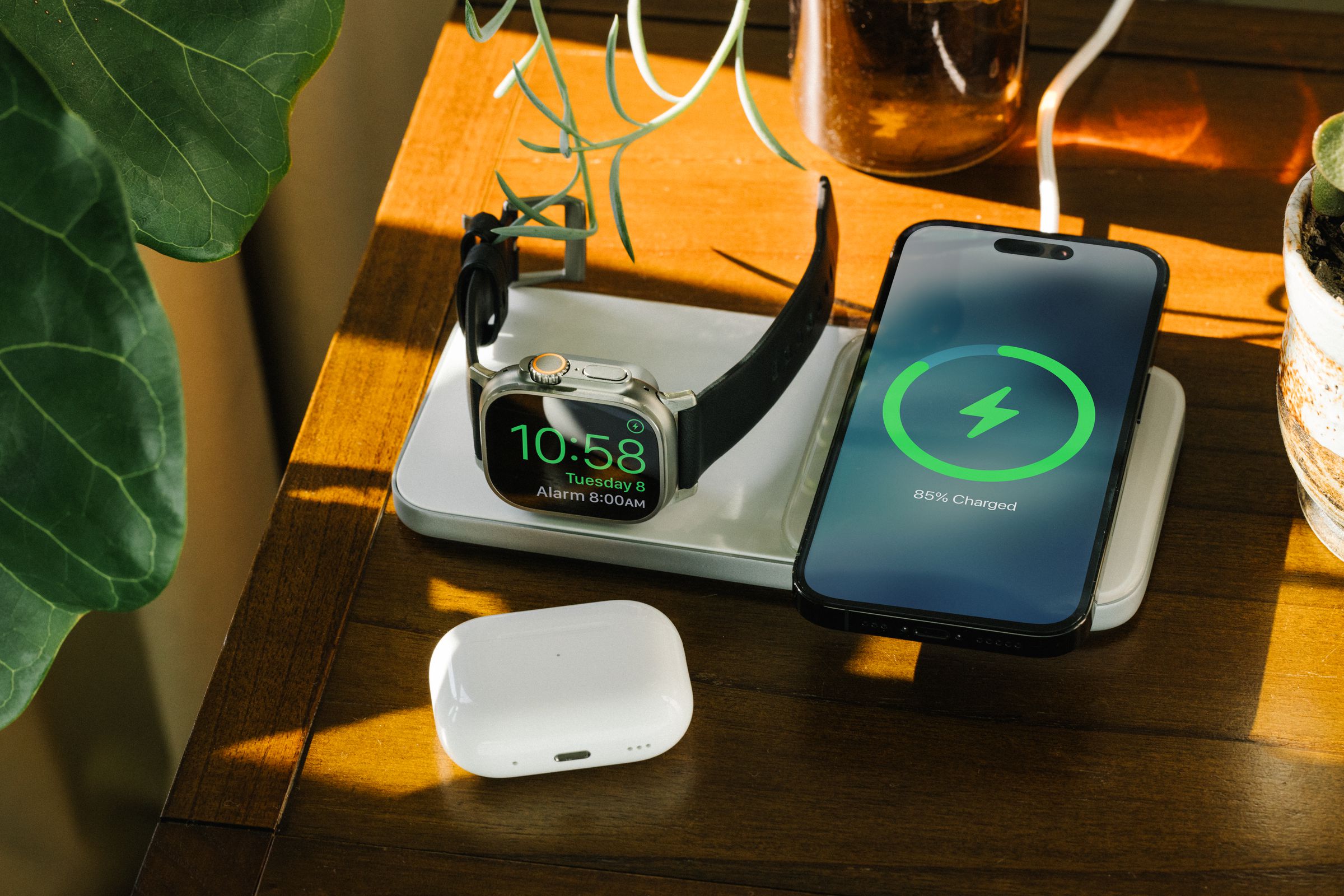 An iPhone and Apple Watch on a table charging on Nomad’s Base One Max wireless charger, with a set of AirPods near.