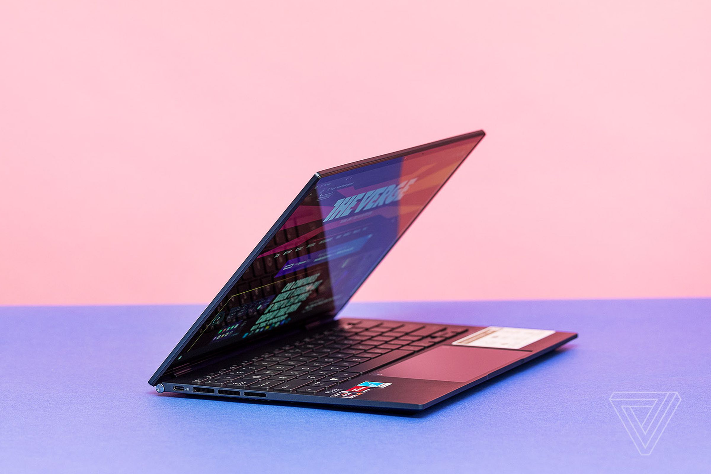 The Asus Zenbook S 13 OLED half open on a pink and blue background. The screen displays The Verge homepage.