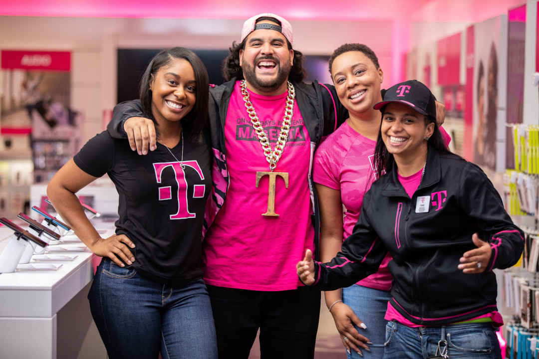 Four people in all races wearing T-Mobile uniform in hugging and smiling inside a T-Mobile store