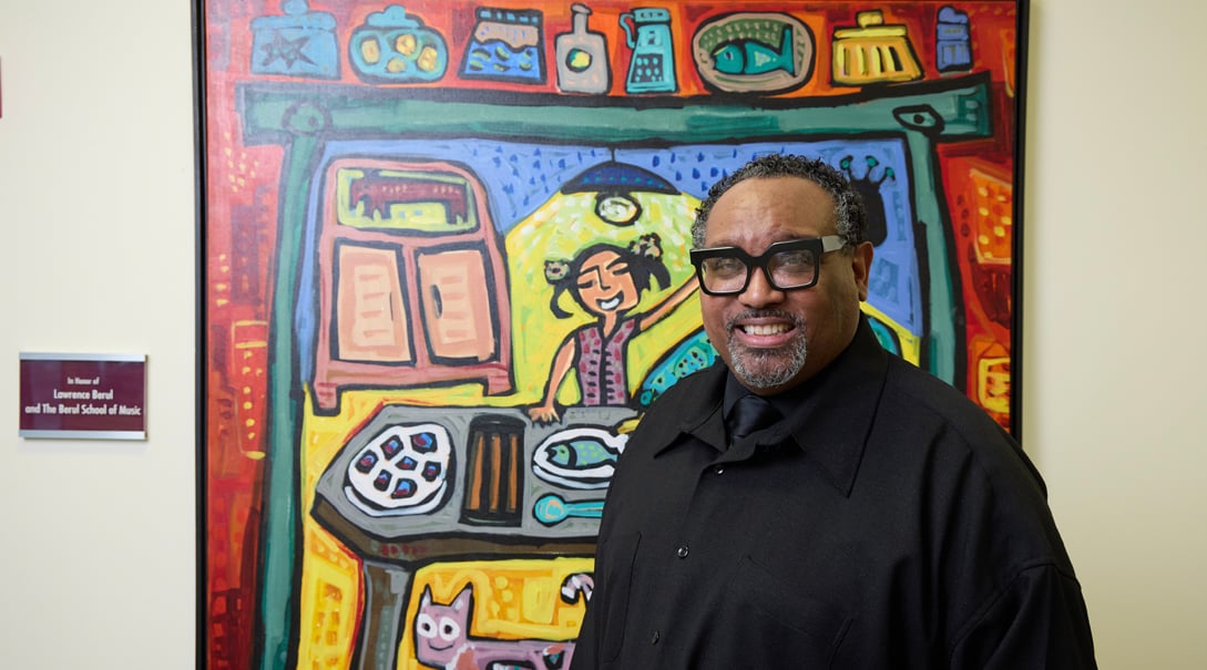 Artist-in-residence, Greg Scott, dressed in black and smiling while standing in front of painting.