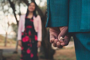 person holding a diamond ring to their side, with a smiling woman out of focus in the background