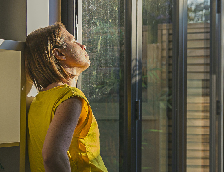 File image of a woman leaning on a bookshelf by a window with her face bathed in sunlight.