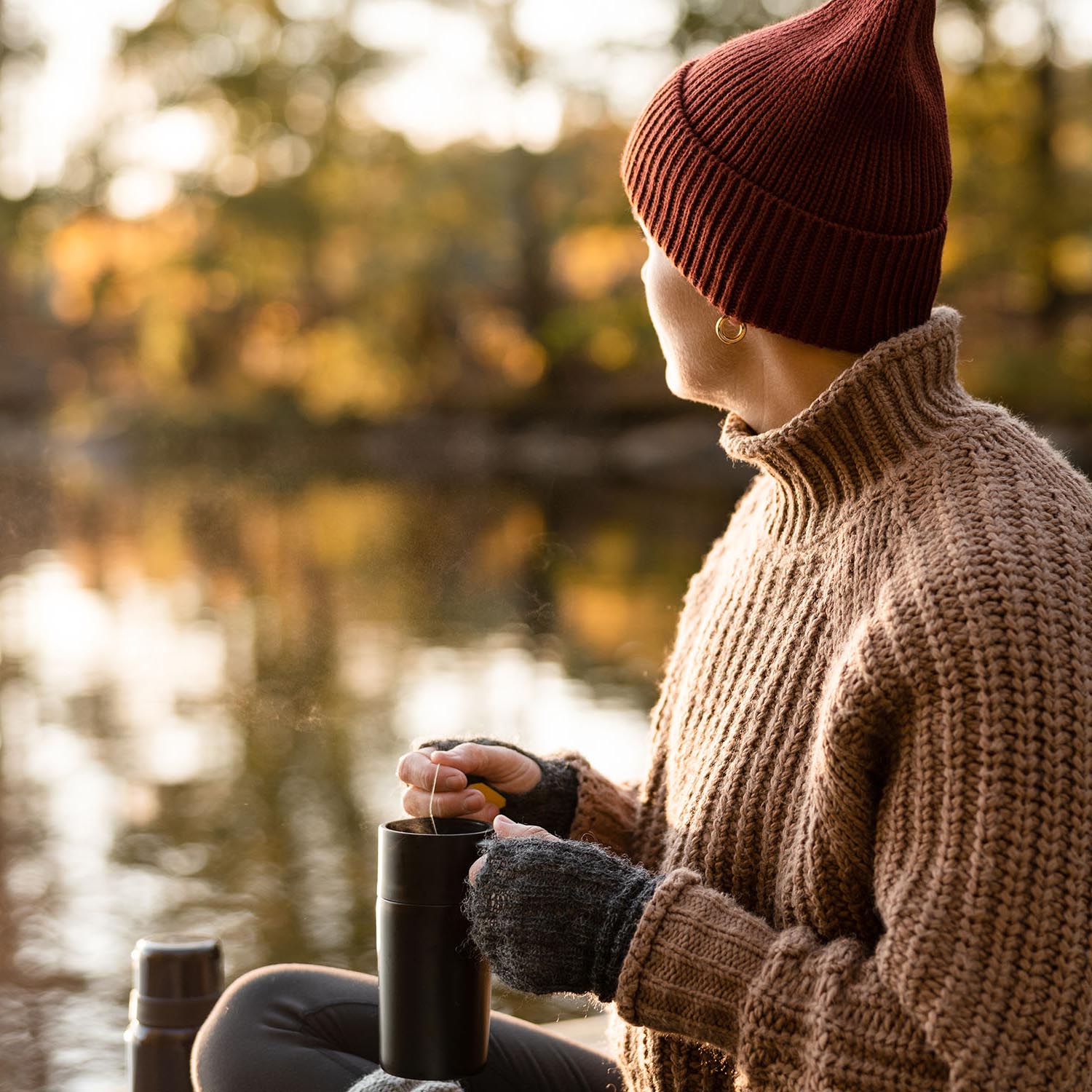 A woman preparing and drinking some fresh hot tea in a travel mug. She is sitting on a small jetty on the lake in autumn.