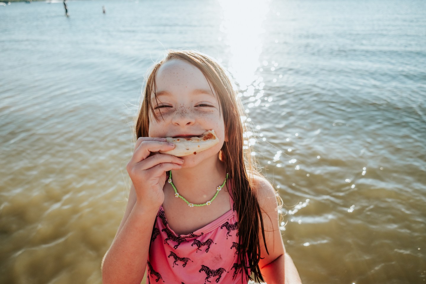 Young girl in a pink swimsuit standing in shallow water eating a snack