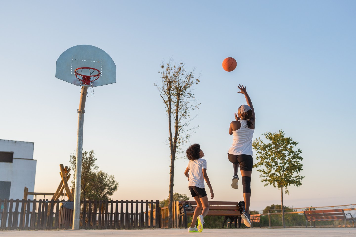 Mother shoots the ball into the basketball basket on a sports court in a park at dusk while her son looks on, we see them from behind