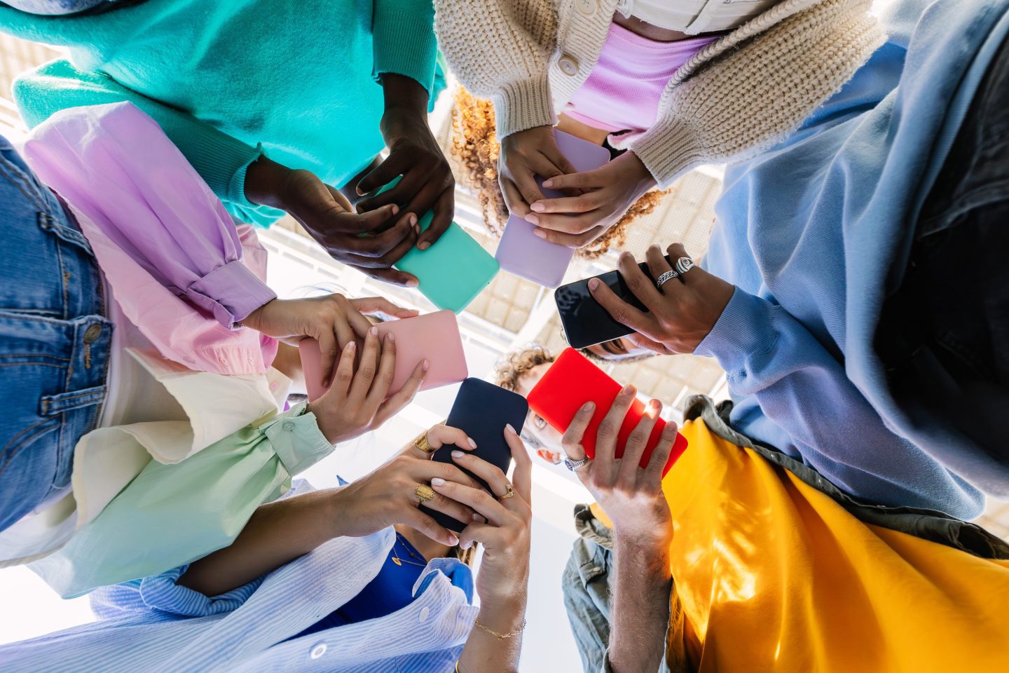 Diverse group of young people standing in a circle, all looking at their phones, shot from below their hands