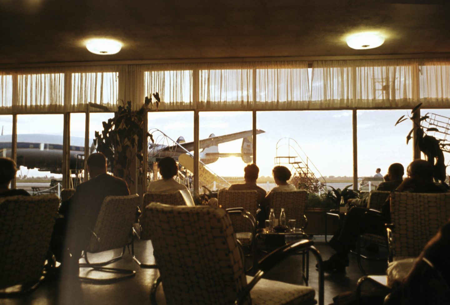 Passengers seated in a vintage airport departure lounge, gazing through the window at an airplane parked at the gate