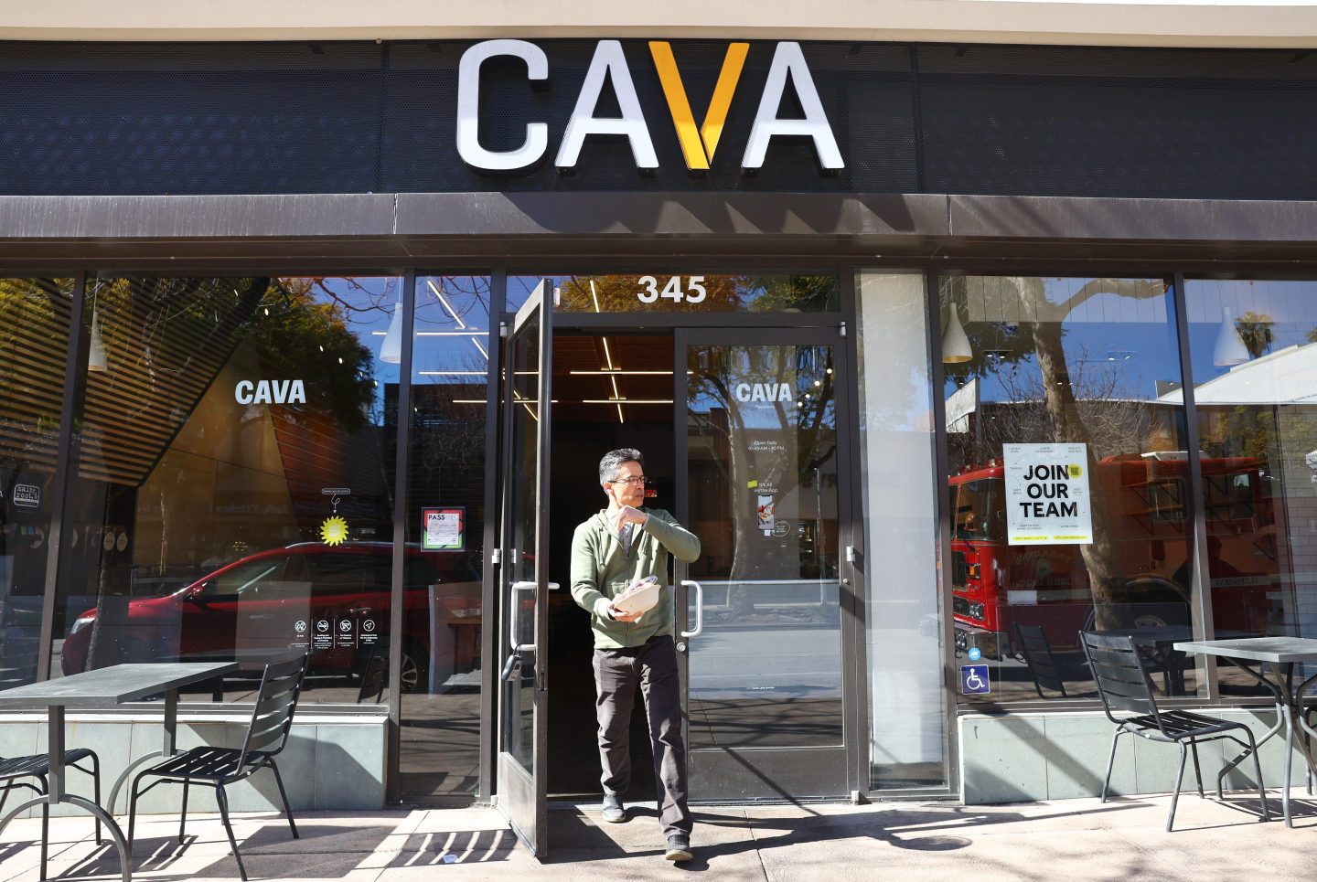 A man in a green sweater and brown pants carries his food through the door of a Cava restaurant.