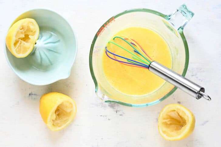 A large liquid measuring cup with a mixture of raw eggs and lemon juice with a whisk in it, with lemon halves and lemon juicer next to it.