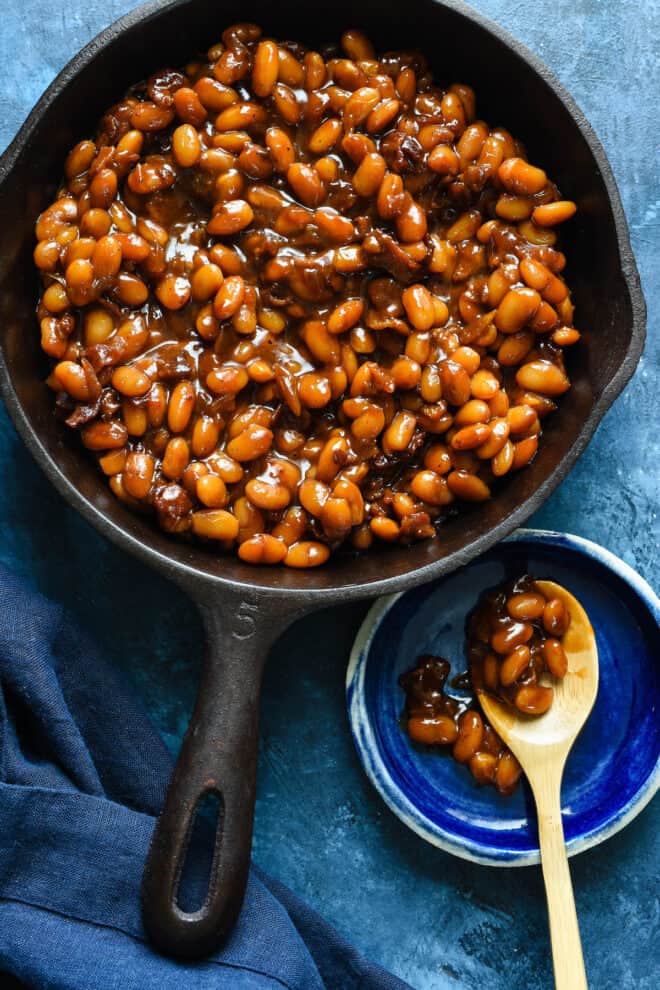 Cast iron skillet filled with glazed Great Northern beans with bacon, with wooden serving spoon resting on small blue dish next to skillet.