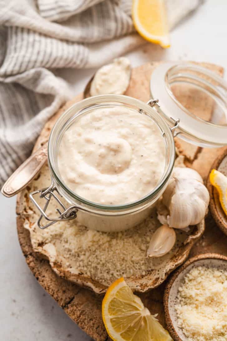 A lidded glass jar filled with homemade Caesar dressing, surrounded by the ingredients for the dressing.