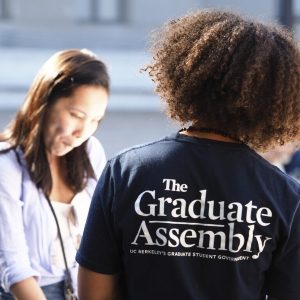 A graduate assembly representative wearing a t-shirt that reads 'The Graduate Assembly' on the back faces another student.