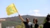 A man waves the Hezbollah flag in the southern Lebanese village of Aita al-Shaab, which has come under Israeli fire numerous times, June 29, 2024. An Israeli military position can be seen in the background.