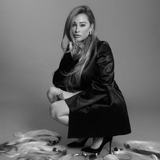 A woman with hair extensions poses in black and white, surrounded by an array of stylish shoes.