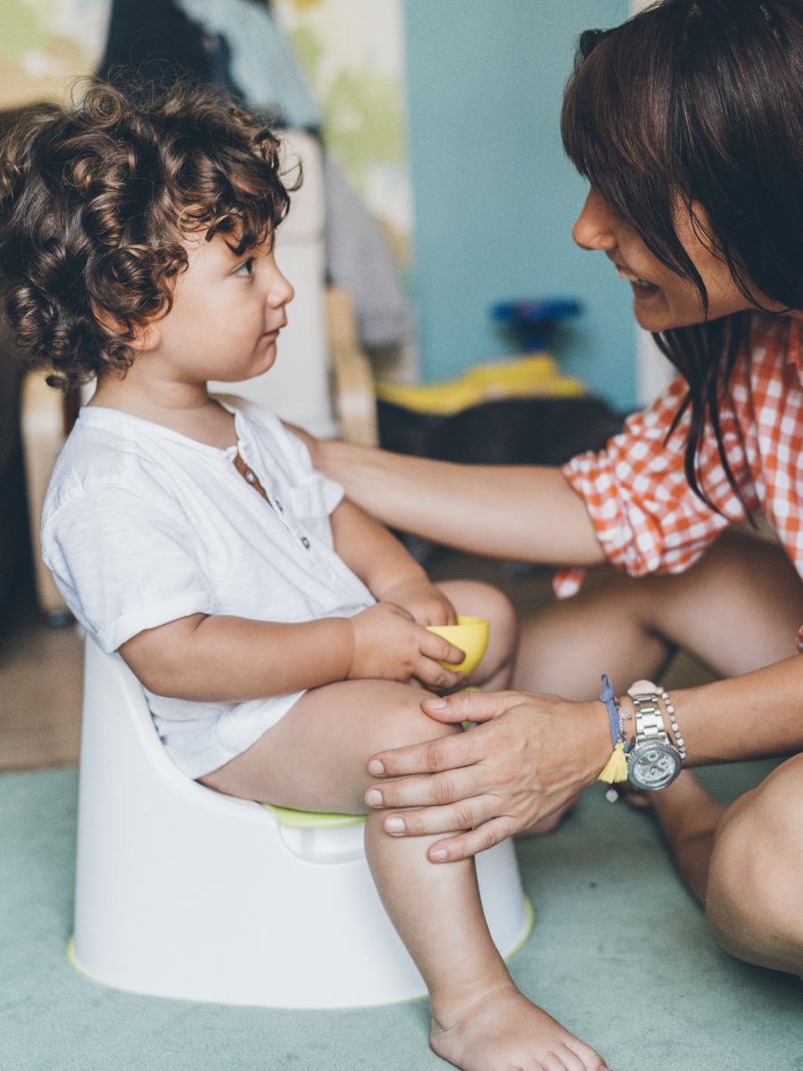 toddler with curly brown hair sits on training potty with supportive mother