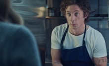 Carmy from "The Bear" in a restaurant kitchen, wearing a blue apron and white t-shirt.