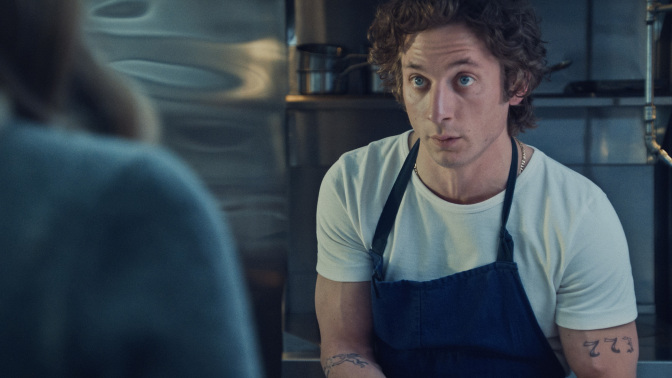 Carmy from "The Bear" in a restaurant kitchen, wearing a blue apron and white t-shirt.