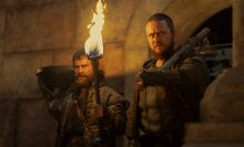 A man with a beard and a larger man stand in a castle holding a fiery torch.