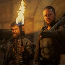 A man with a beard and a larger man stand in a castle holding a fiery torch.