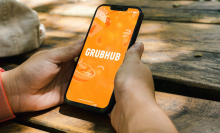 Girl in the park holding a smartphone with GrubHub food ordering and deliveryapp on the screen. Rustic wooden table. Rio de Janeiro, RJ, Brazil. July 2022.