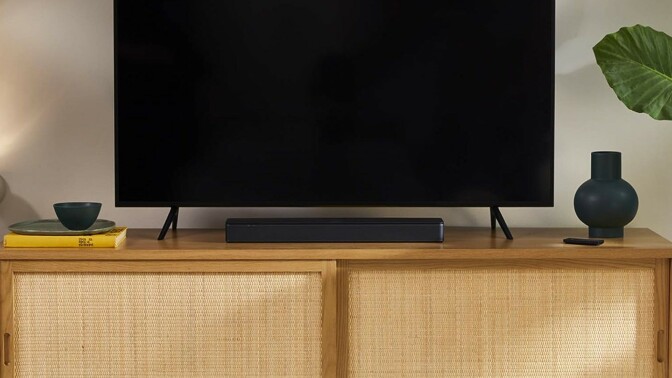 a bose tv speaker soundbar sits below a large TV screen, on top of a light brown entertainment center. The room also has a green plant and the entertainment center has a vase and a stack of books on either side of the TV