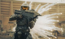 A still from the "Halo" TV series. Master Chief aims a machine gun at a target that's out of view as something explodes behind him.