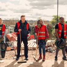 Six people (the cast of "The Outlaws) wearing red community service vests and standing in a line while holding tools. 