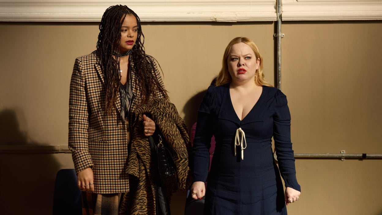 Two women dressed in stylish clothes look concerned against a wall.