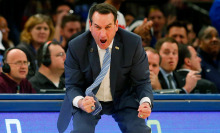 Duke head coach Mike Krzyzewski of the Duke Blue Devils leans forward, apparently shouting, with fists clenched. 