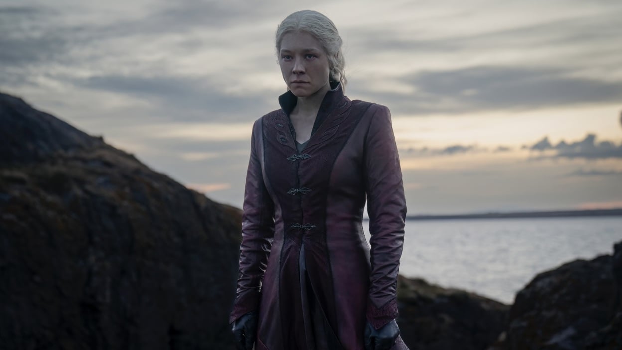 Rhaenyra from "House of the Dragon" stands on a rocky cliff by the sea, wearing a long red coat.