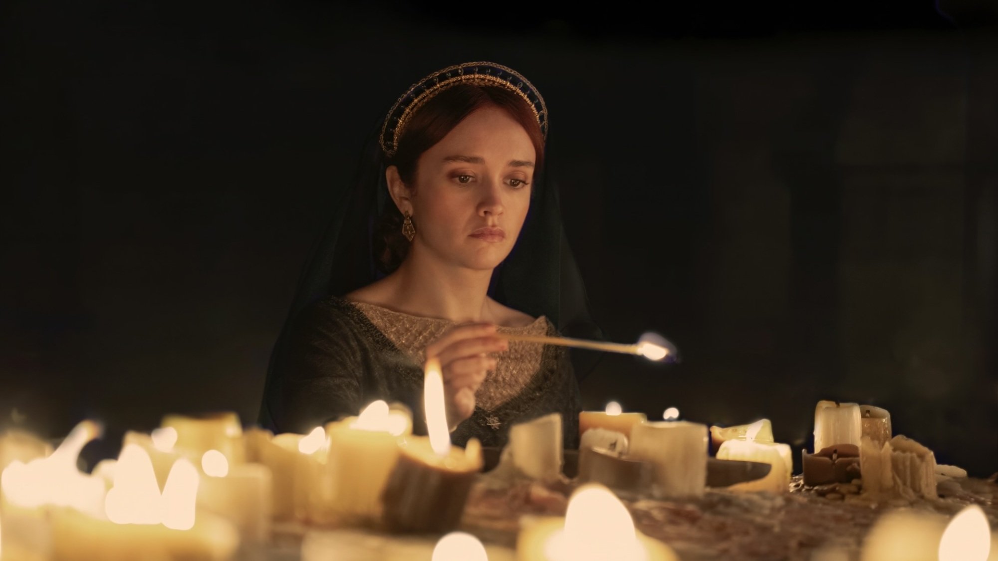 Alicent from "House of the Dragon" lights candles at an altar, wearing a green dress and black veiled headdress.