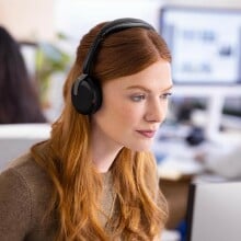 a person with red hair wears the  Jabra Evolve2 65 headset while looking at a computer screen. The setting it an office environment with other people working in the background at computer stations.