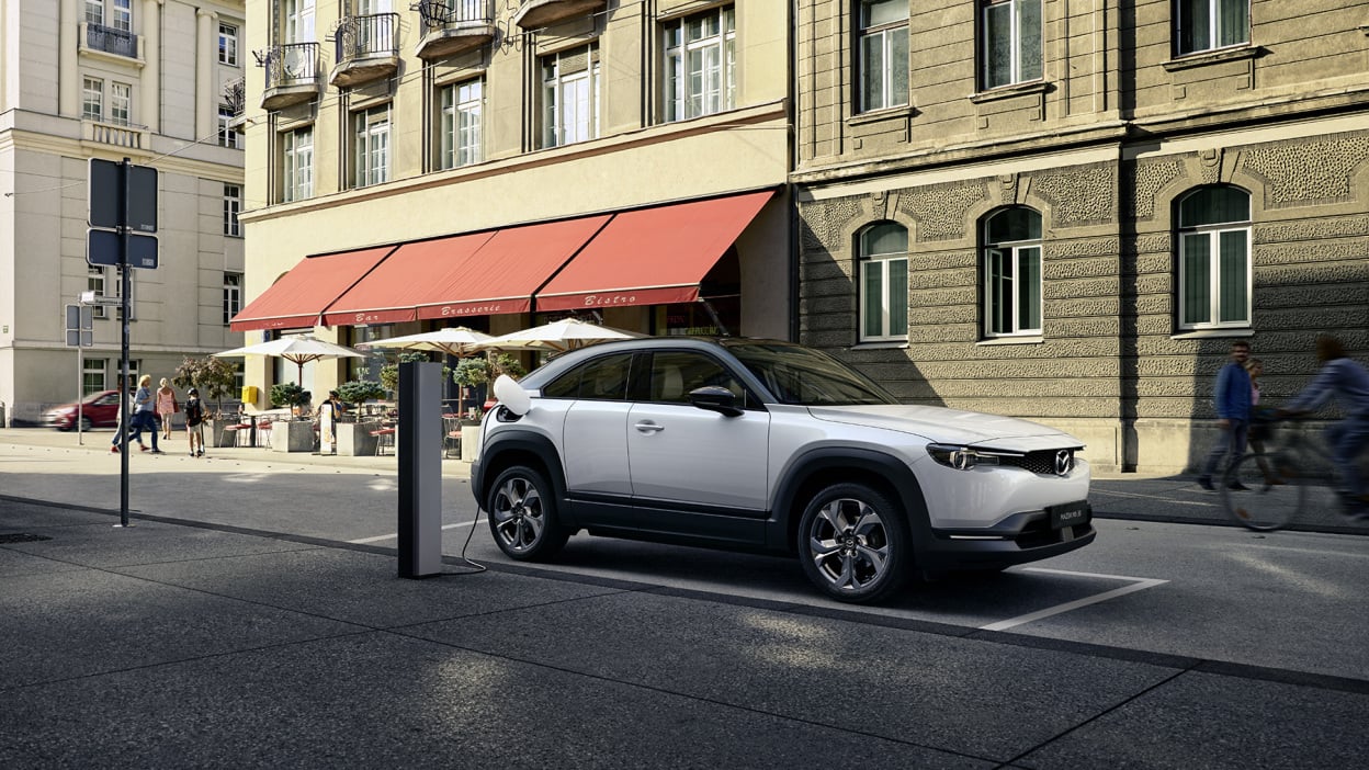 A white SUV plugged into a charging station on a street with a biker and a red awning.