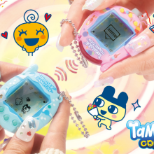 Two pairs of white hands each hold a Tamagotchi Connection, one in blue and one in pink.