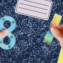 A pair of hands grasp and point to colorful stickers on top of a composition notebook.