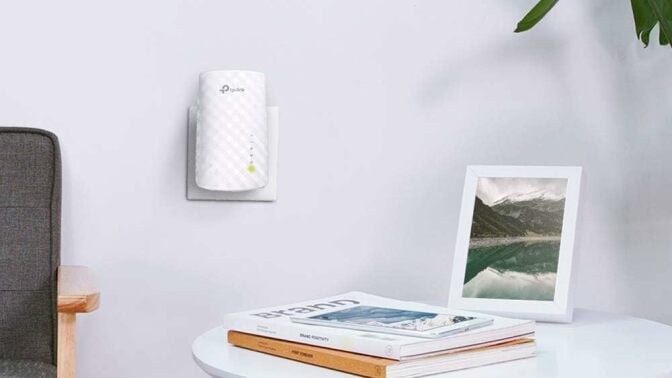 A TP Link WiFi extender is plugged into an outlet on a white wall with a side table and a chair next to it