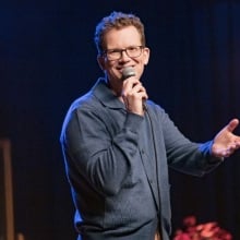 Hank Green performs stand-up onstage in a blue button-up sweater.