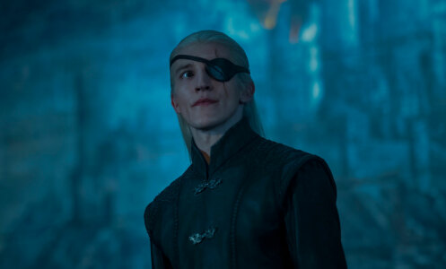 A man with blonde hair and an eyepatch stands in a gloomy room.