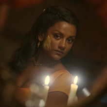 An Indian woman with turmeric paste on one cheek, looking at her reflection in a mirror by candlelight.