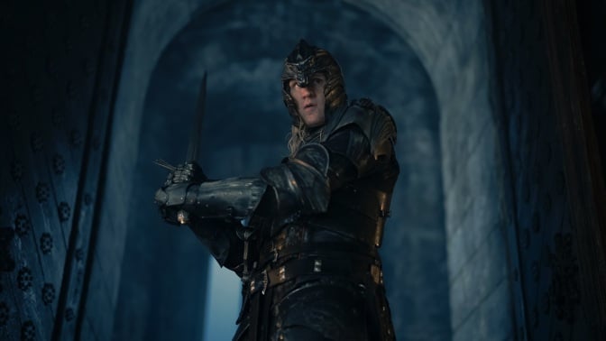 Daemon Targaryen walks through a large doorway in Harrenhal, holding a sword and wearing full armor.