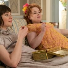 Two young women in Regency attire lounge on a couch eating treats. 