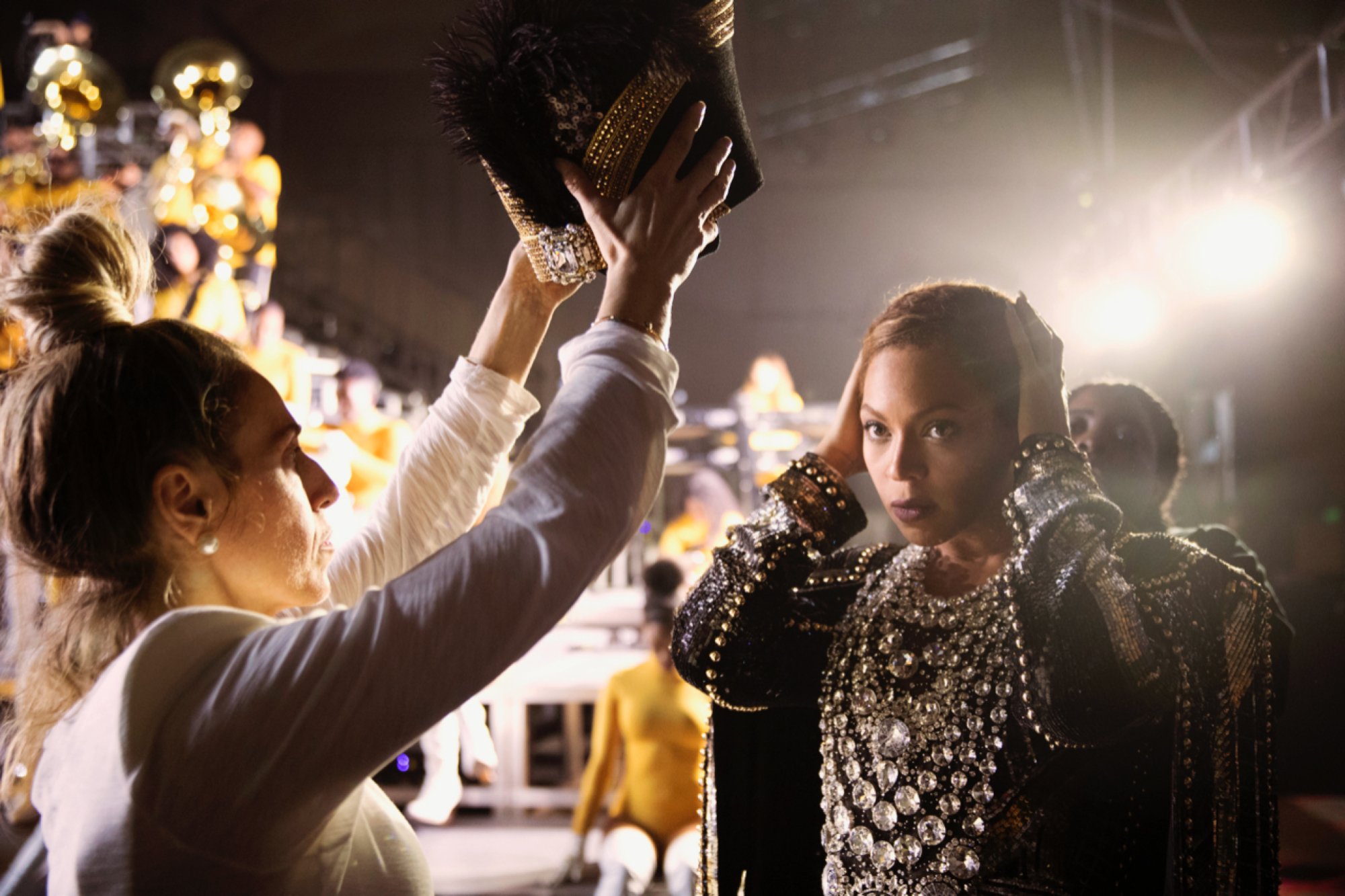 Beyoncé getting ready backstage, adjusting her hair as an assistant offers her a headpiece.
