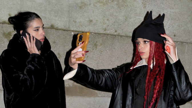 Two young women on a sidewalk, one talking on her cell phone, the other taking a selfie while wearing a black hat with cat ears