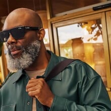 a person wearing the Carrera Smart Glasses with Alexa sunglasses holds a brown leather bag over their left shoulder while looking forward. the person has a salt and pepper beard and a bald head 
