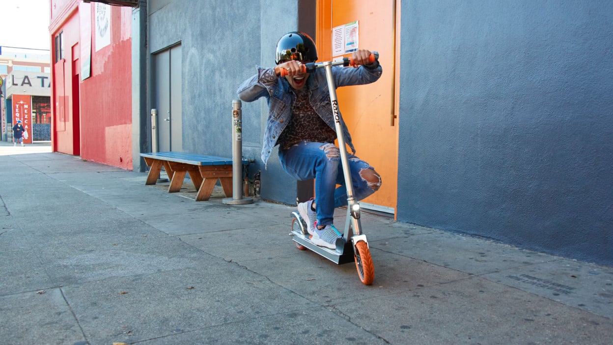 A helmeted person crouch on a scooter in front of colorful garages.