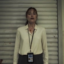 A woman in a blood-spattered white button-up shirt presses her back up against the wall of an interrogation room.