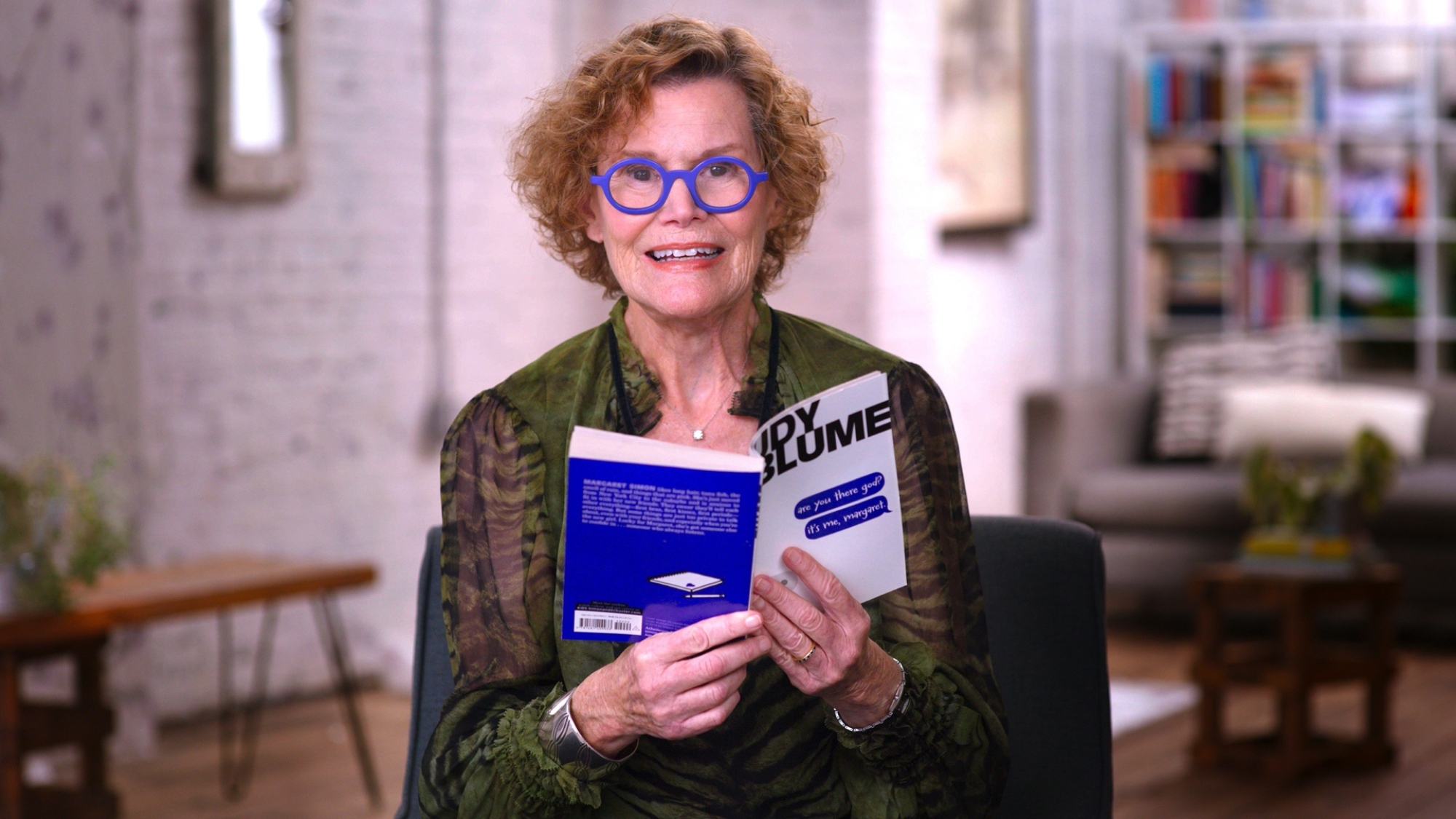 A woman with short curly hair and bright blue glasses is holding a copy of a book called "Are You There, God? It's Me, Margaret."