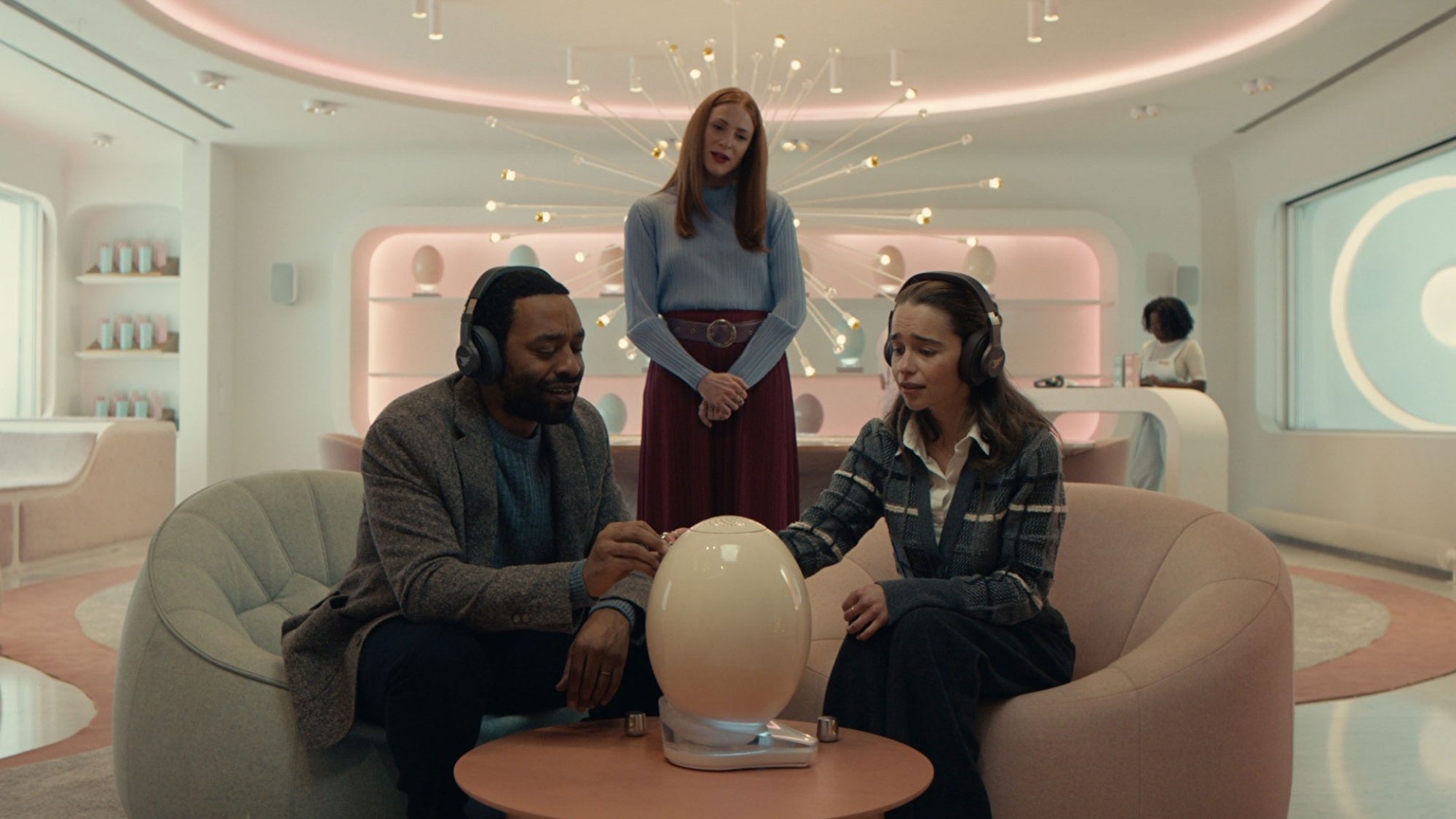 A man and a woman "bond" with a strange egg resting on a table while a woman stands behind them, watching.